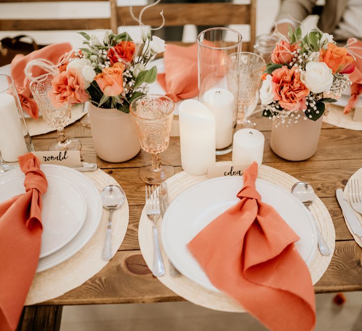 Place settings with coral napkins, coloured glassware and flowers in beige jars. 