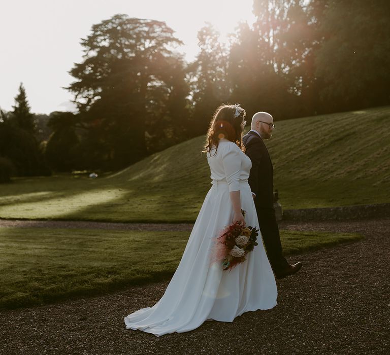 Groom in navy suit and yellow tie walks with bride in white cat eye glasses and bridal headband holding white and pink rose and pampas grass bouquet