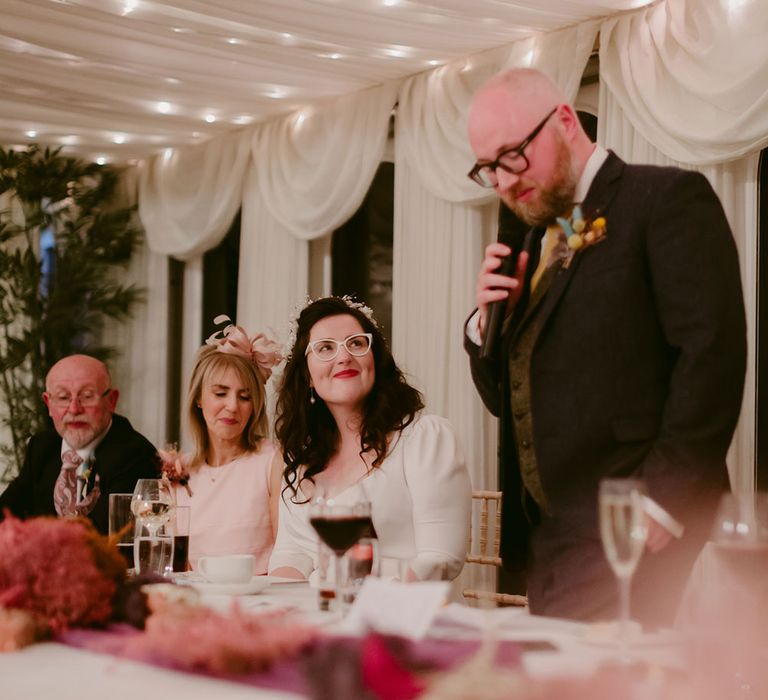 Smiling bride in white cat eye glasses, bridal headband and Rime Arodaky dress smiles at groom in navy suit and yellow tie as he makes speech