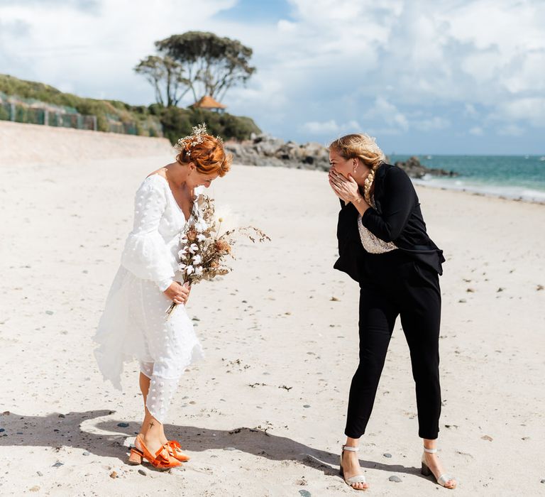 Bride in dress standing behind as bride in suit smiles looking shocked on beach for first look shot