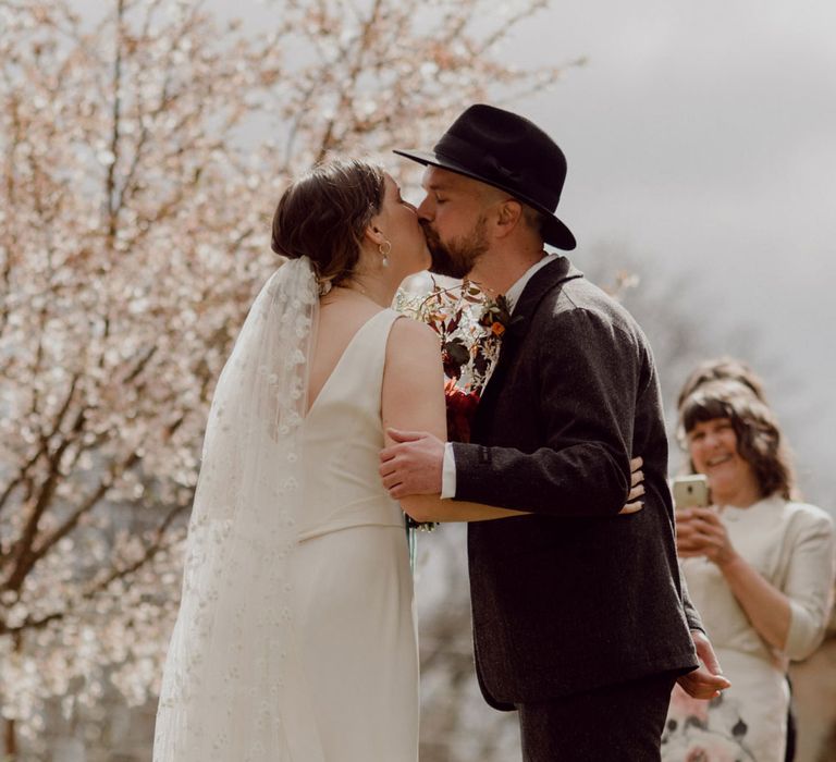 Bride in Charlie Brear wedding dress and daisy chapel length applique veil kisses groom in grey tweed suit and black fedora at garden party wedding at Hayne, Devon