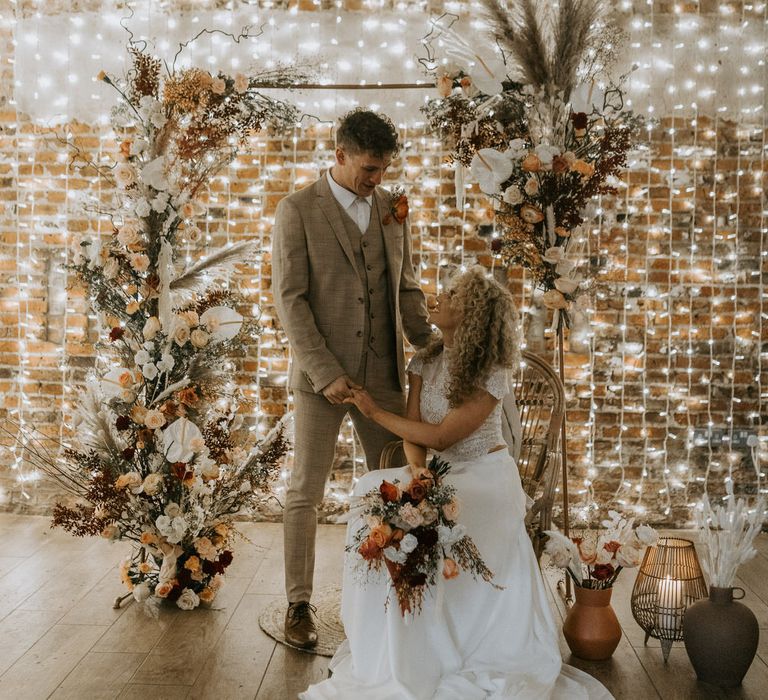 Bride and groom at wedding altar with rattan chair, pampas grass and rose flowers, and a wall of fairy lights