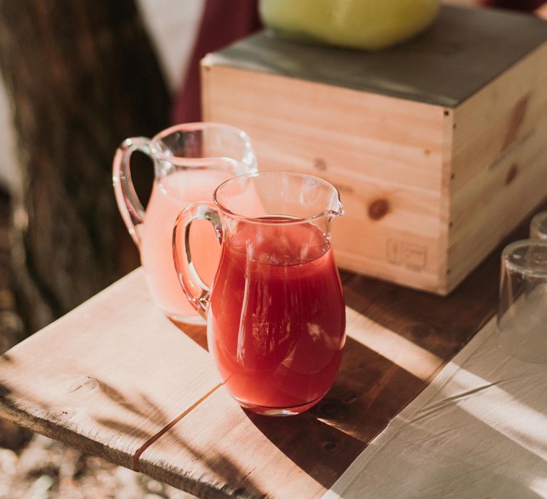 Red & pastel pink drinks in jugs filled with ice sit on wooden tabletop 