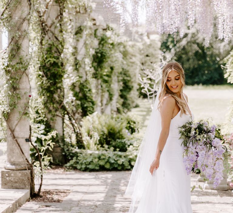 Bride stands beneath pergola with hanging wisteria and lilac florals above her