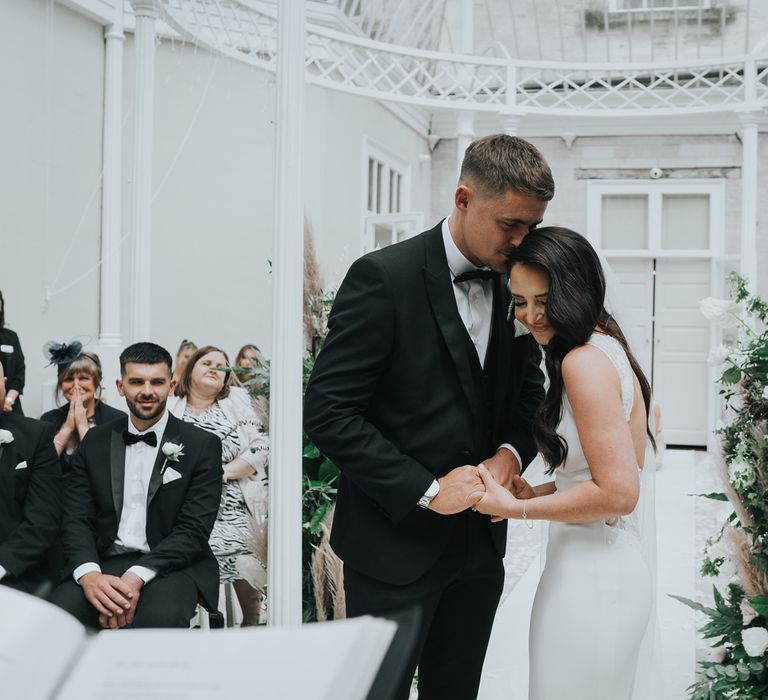 Groom in black tuxedo and bride in white Pronovias wedding dress smile and hold hands during wedding ceremony in the glass conservatory at Came House Dorset