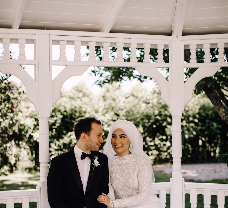 Bride and groom seated in a white gazebo. Groom is in black tie and bride wears high neck and long sleeve 3D lace gow