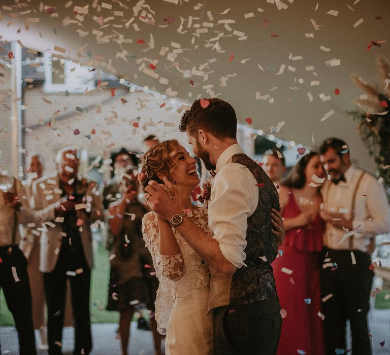 Bride in white Rime Arodaky wedding dress and mesh floral embroidered top dances with groom in white shirt and light brown waistcoat in fairy lit tipi whilst confetti falls at garden wedding with burnt orange wedding theme