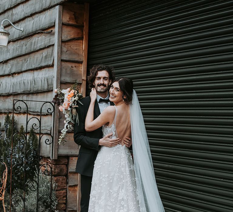 Groom in a black tuxedo and bow tie embracing his bride in an appliqué Made With Love wedding dress and cathedral length veil 