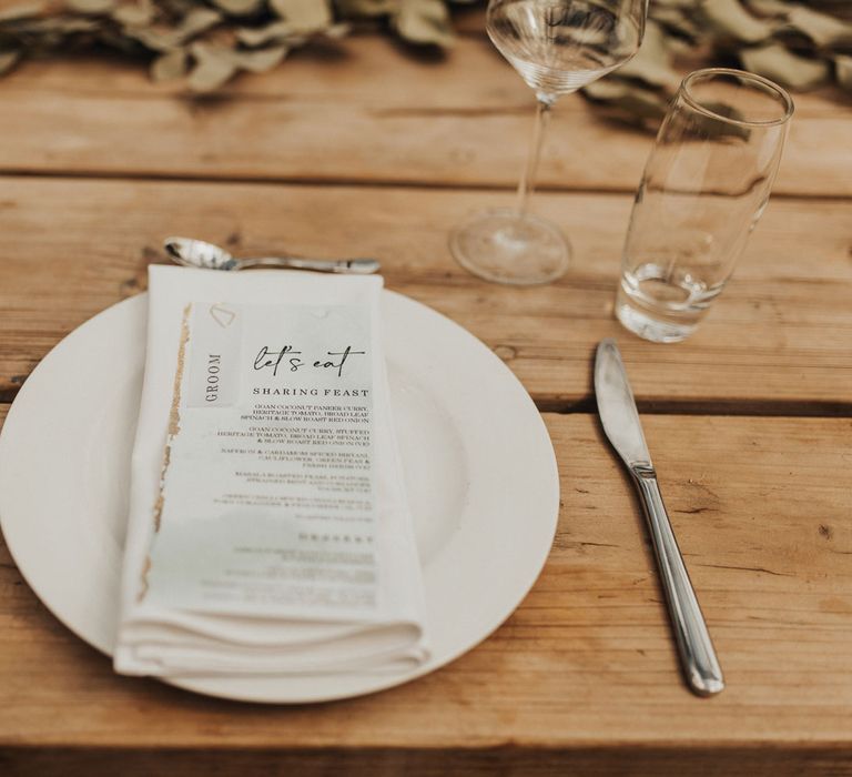 White plate setting with napkin and menu on wooden table set with cutlery and glassware with foliage table garland for wedding breakfast at Anran Devon