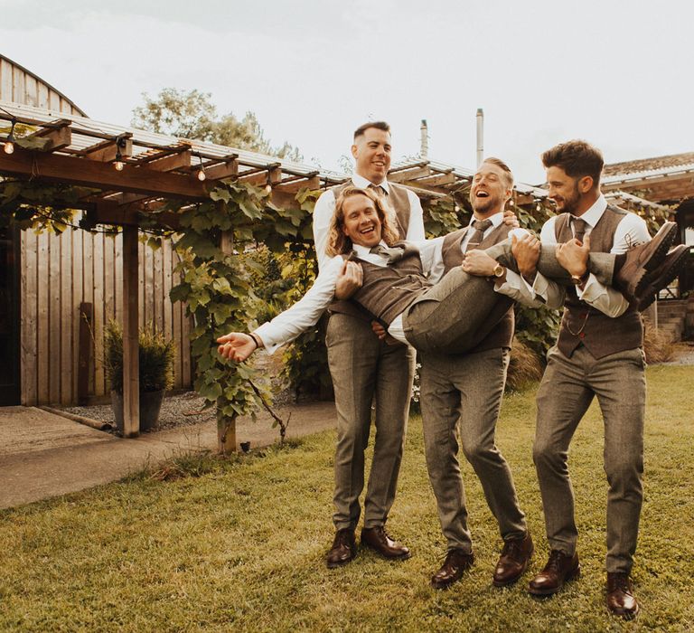 Three groomsmen in sage green trousers, waistcoats and ties hold groom in sage green trousers, waistcoat and tie in the garden at Anran Devon after wedding