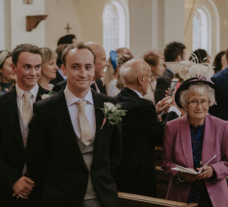 Groom in black morning coat, olive green waistcoat and bespoke olive leaf print tie waits for bride at the end of the aisle during church wedding ceremony in Surrey