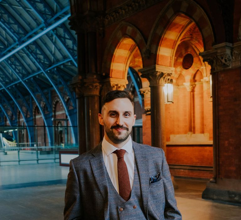 Groom poses for photo whilst wearing burgundy tie and light grey suit with waistcoat on his wedding day