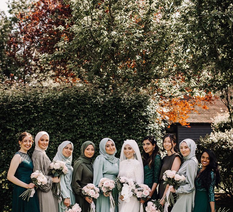 A Muslim bride stands with her bridal party for wedding photo. She wears white and they all wear varying hues of green and grey.