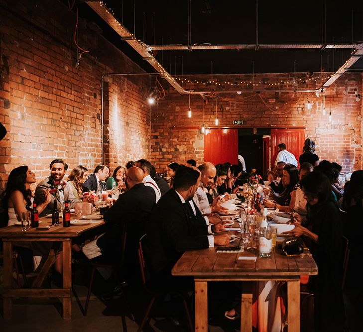 Wedding guests sit at long wooden tables for a wedding breakfast.