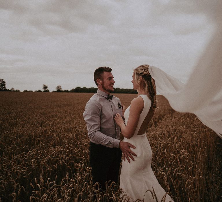 Brides veil blows in the wind as her groom wraps his arms around her whilst stood in golden field