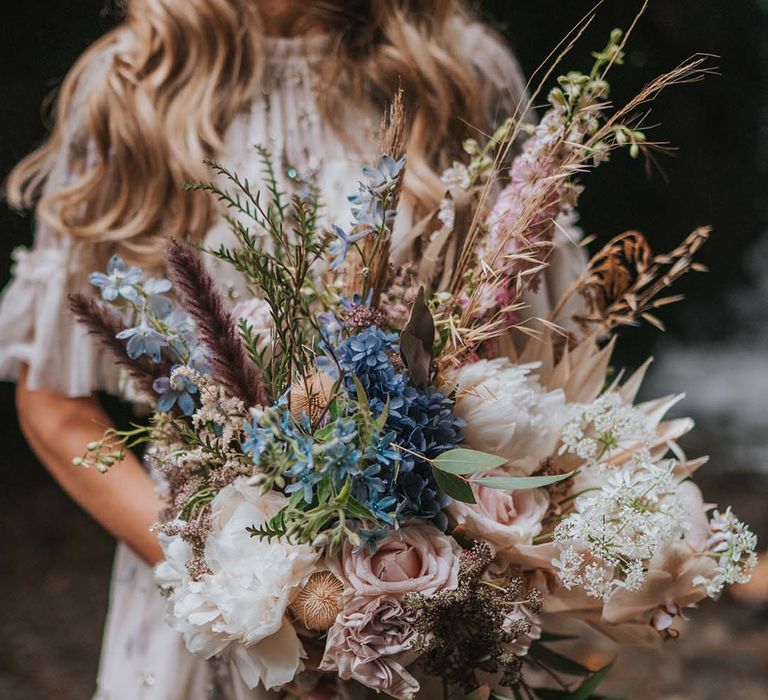 Bride holds out floral bouquet complete with pastel pink roses and pale blue florals 