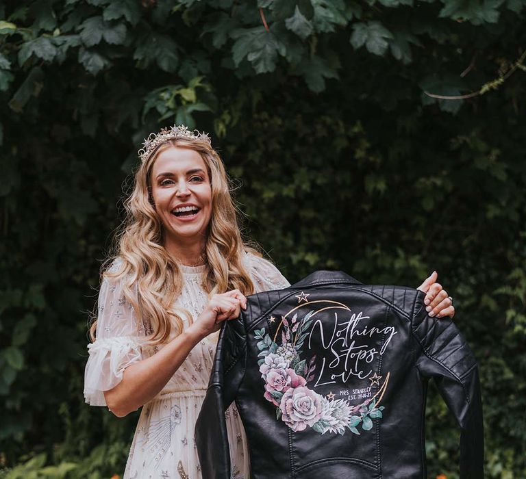 Bride holds up her personalised leather jacket complete with name and wedding date and floral illustrations 