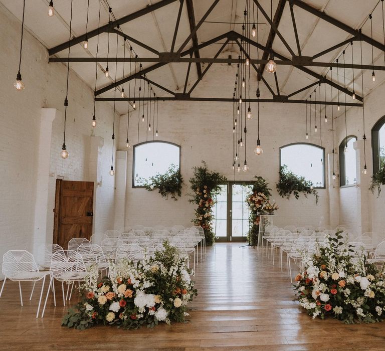 Wedding ceremony room at Osea Island with hanging festoon lights, orange and peach wedding flowers and white metal chairs 