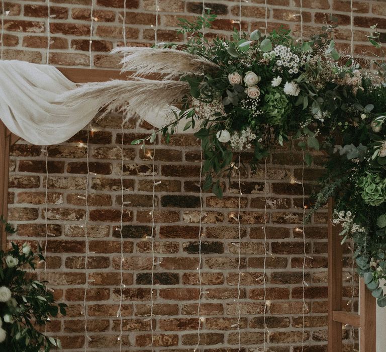 Flower cloud on wooden wedding arch with drapery in front of fairy string lights for Isle fo Wight wedding with macrame wedding decor