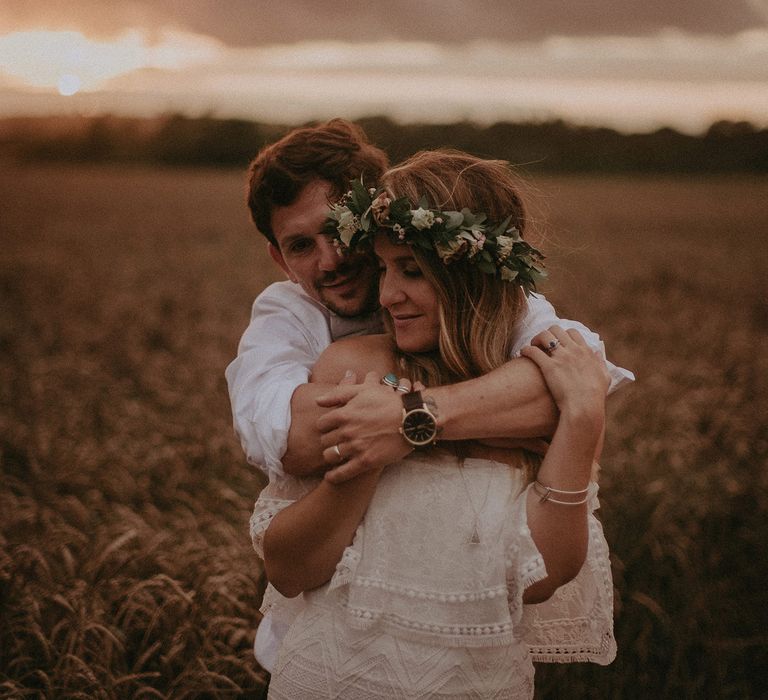 Groom in white shirt hugs bride in Grace Loves Lace bardot wedding dress and flower crown as they stand by a wheat field at golden hour at Isle of Wight wedding with macrame wedding decor