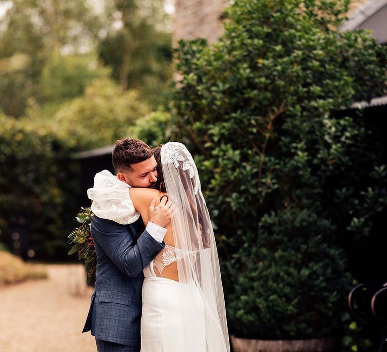 Groom in a blue check suit embracing his bride in a bespoke wedding dress with cut out back, lace puff sleeves and matching veil 