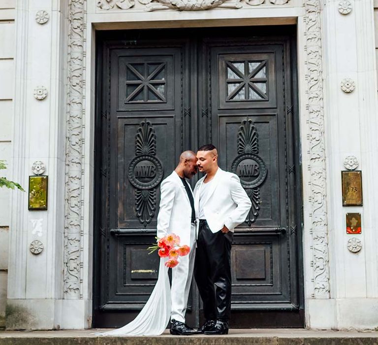 two stylish grooms in a white satin suit with a cape, leather trousers and double breasted jacket embracing at their city elopement 