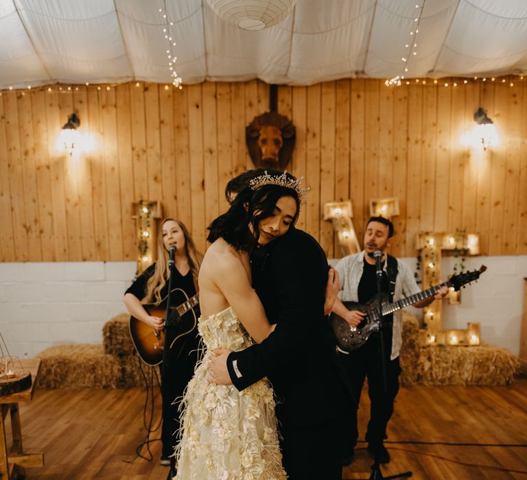 Bride in strapless feathered wedding dress and headpiece hugs groom in dark blue suit as two guitarists sing in the background during rustic wedding ceremony