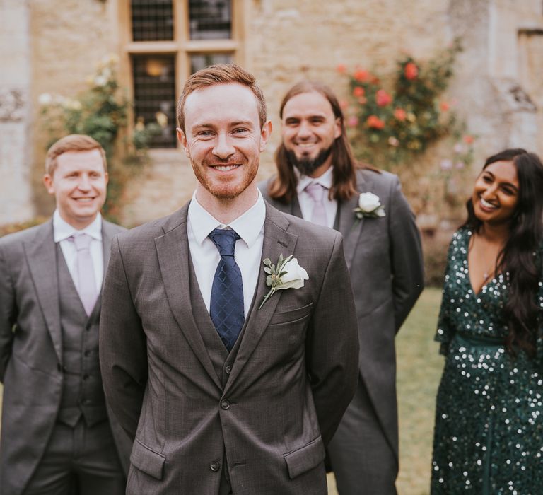 Smiling groom in grey three piece suit with blue tie and white rose buttonhole stands with groomsmen in grey morning suits and woman in emerald green sequin dress for outdoor wedding in Buckinghamshire