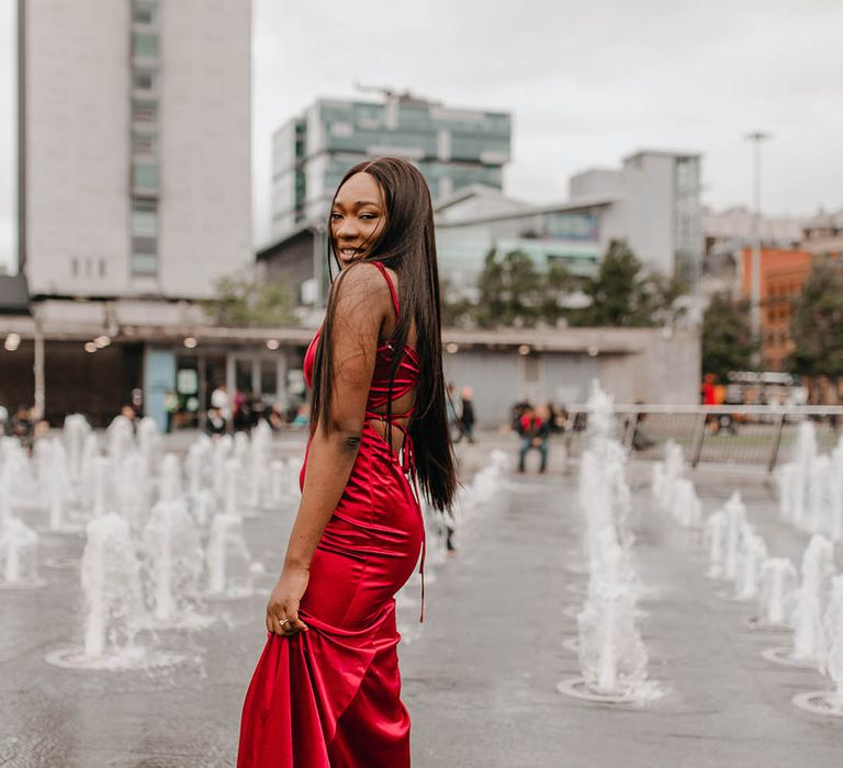 Bride wears sleek red gown as she stands in front of fountains on her wedding day