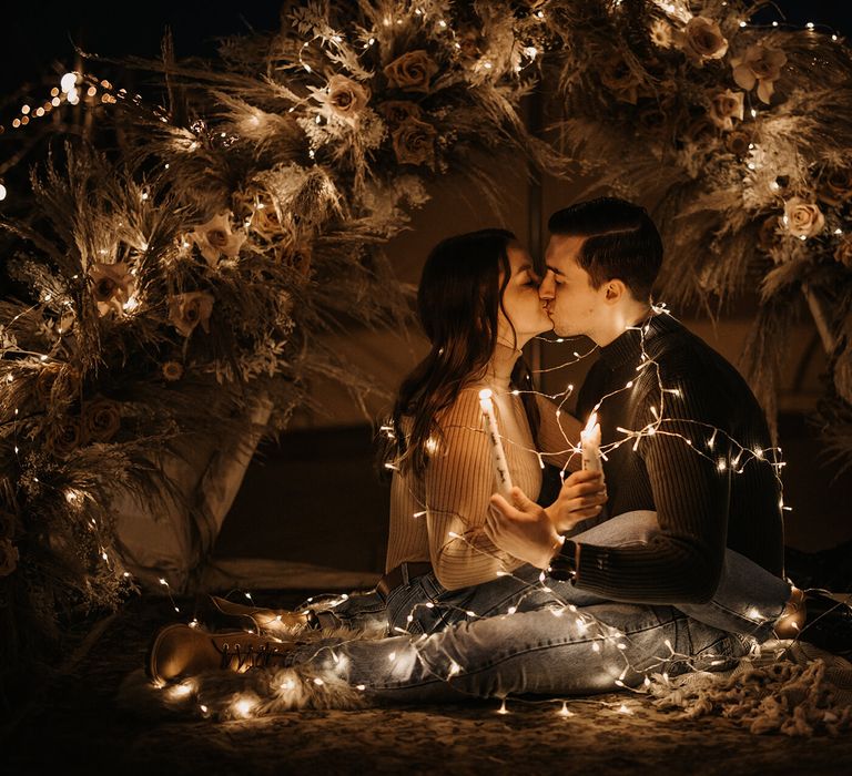 Bride and groom-to-be wrapped in fairy lights at their intimate boho campfire wedding proposal 