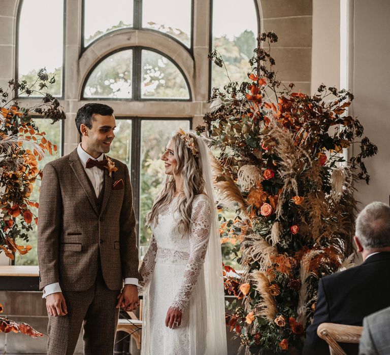 Bride in boho lace two piece wedding dress, veil and dried flower crown smiles at groom in three piece brown tweed suit and bow ties as they stand in wedding ceremony room surrounded by orange and green flower columns