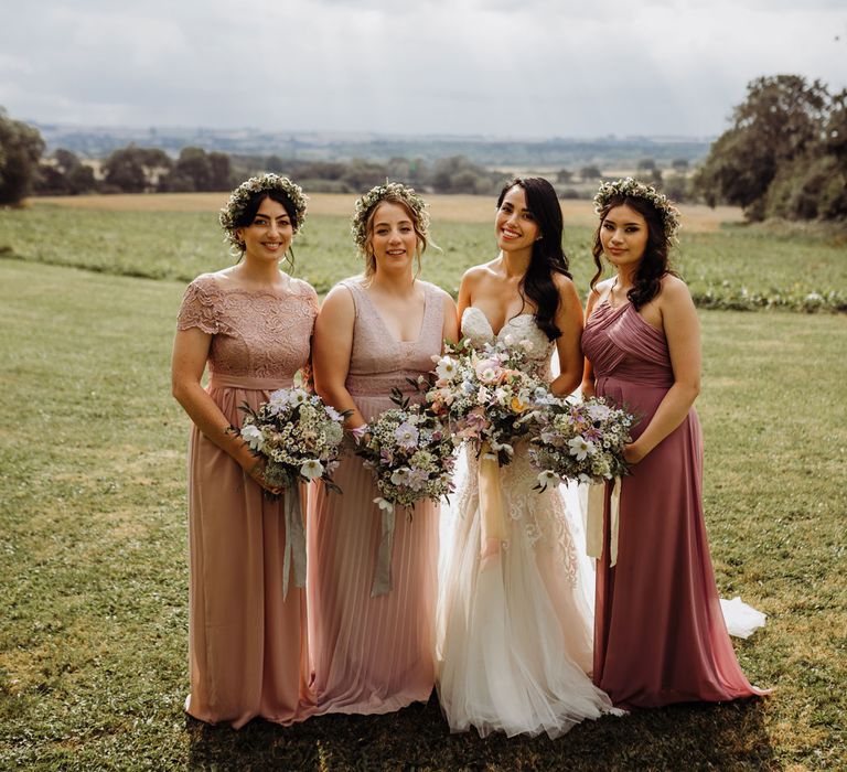 Bride in lace and tulle wedding dress stands with bridesmaids in mixed pink bridesmaid dresses and flower crowns in field holding multicoloured taste bridal bouquets