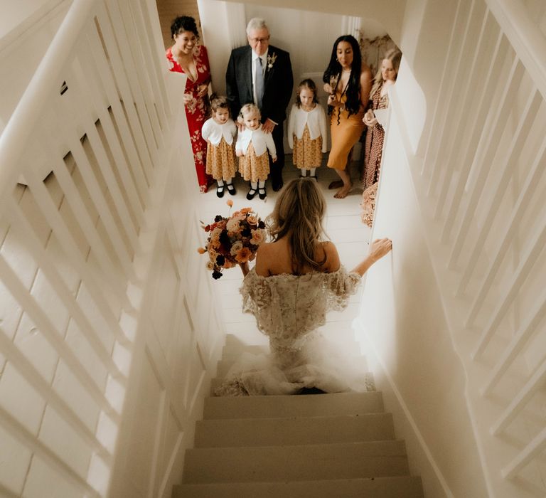 Bride in applique wedding dress holding mixed rustic bridal bouquet walks down stairs towards wedding guests