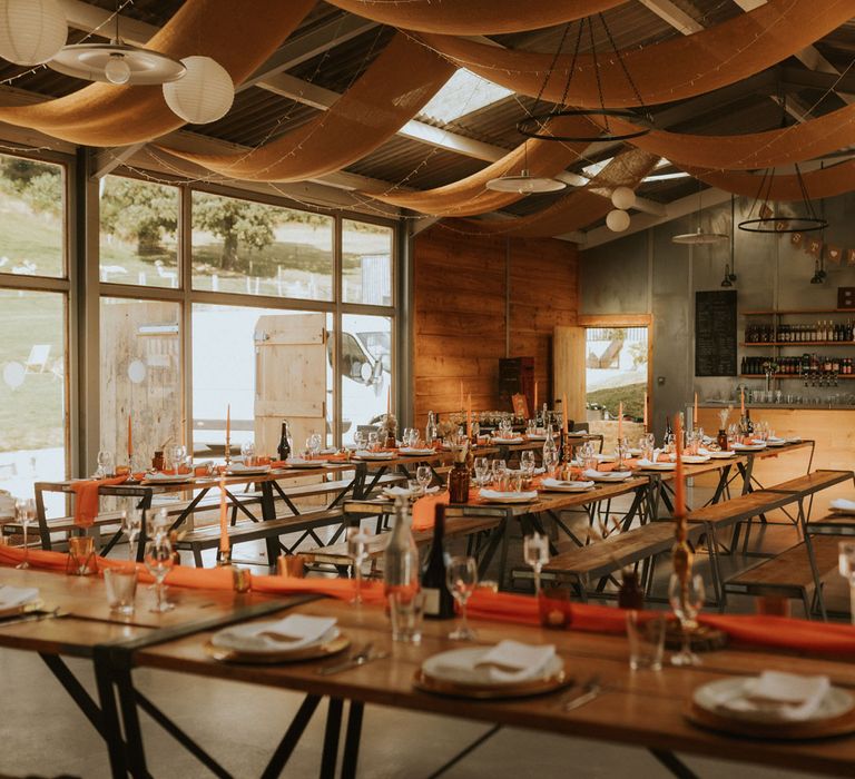 Modern rustic wedding reception in barn with long wooden tables, orange table runners, orange dinner candles and rustic wedding dinnerware