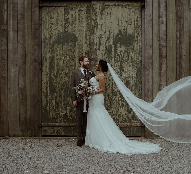 Bride & groom kiss in front of rustic barn door as brides veil blows in the wind