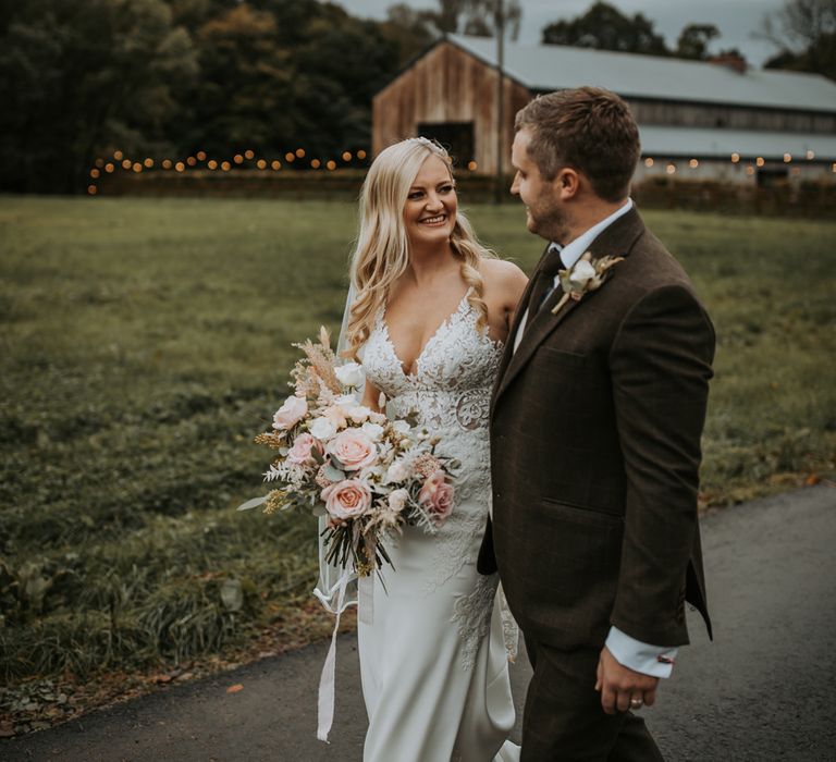 Bride in lace wedding dress and veil holding white, pink and green bridal bouquet smiles as she walks with groom in brown suit outside at Lake District wedding