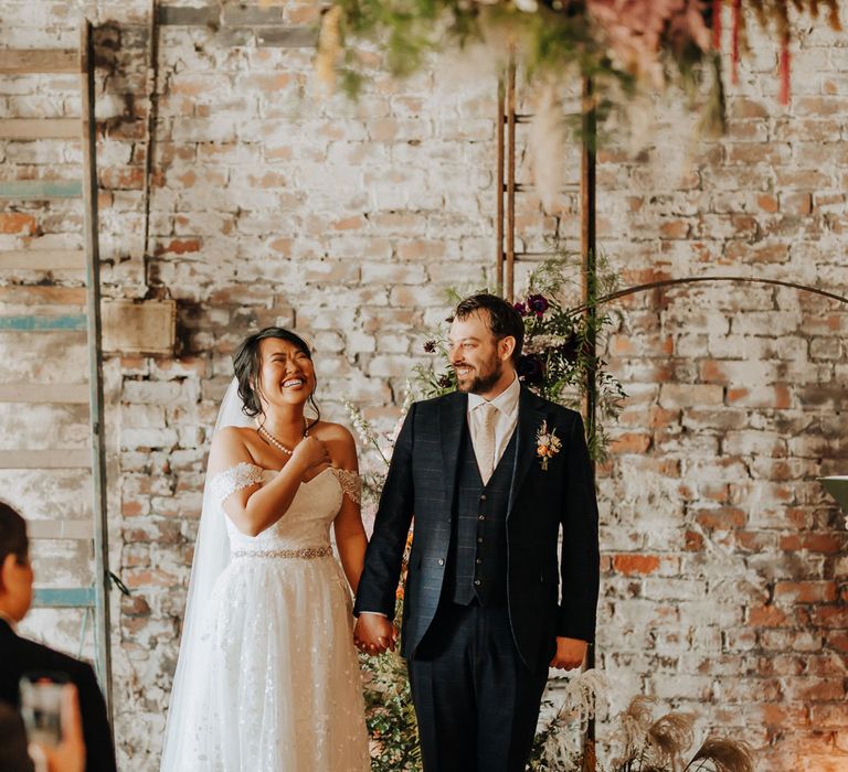 Bride in off the shoulder wedding dress and pearl necklace holds hands with groom in blue check three piece suits and floral buttonhole during wedding ceremony