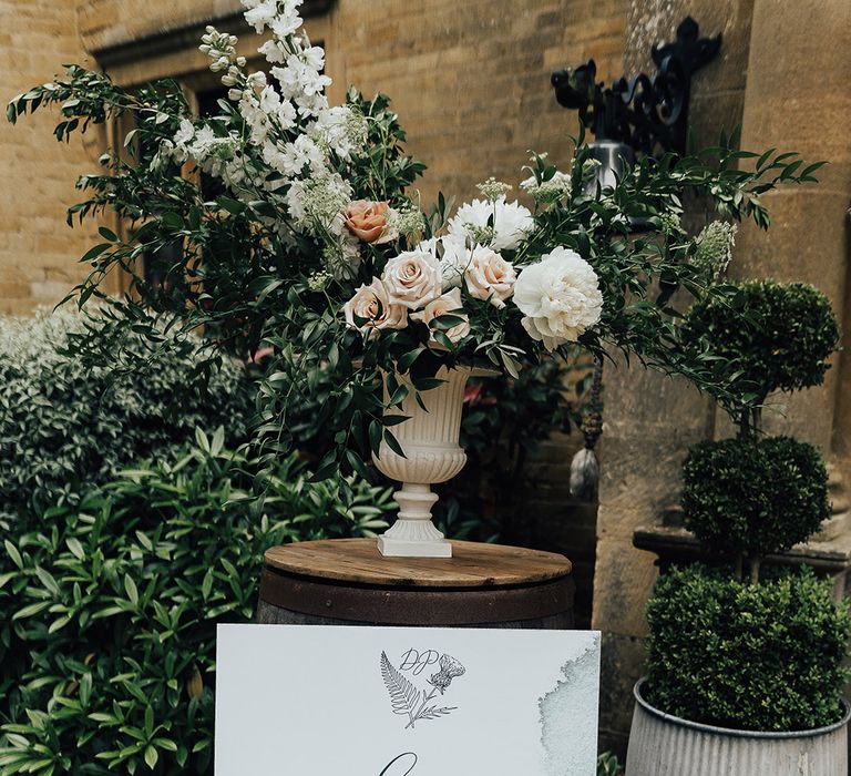 Wedding welcome sign with green watercolour design propped up against a urn floral arrangement with roses 