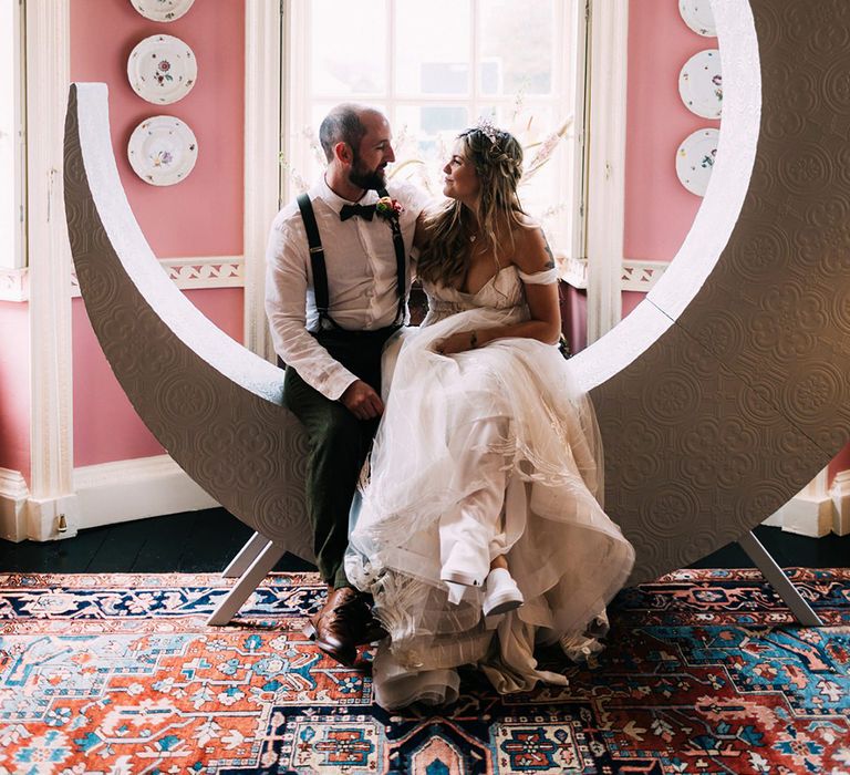 Bride and groom seated on large crescent moon prop at the altar 