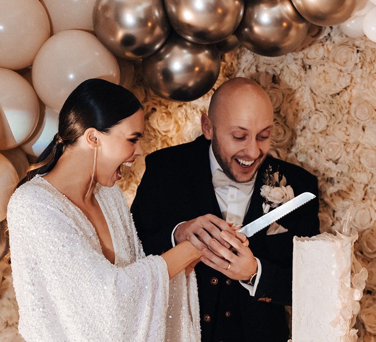 Jessamy Stoddart in a sparkly wedding dress cutting her square wedding cake in front of a flower wall with balloon installation 