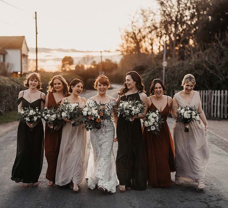 Laughing bride and bridesmaids walk along the road as golden hour hits