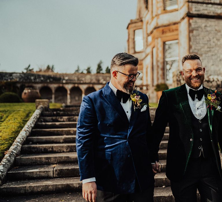 Smiling grooms in green and blue velvet suit jackets smile together around the grounds of the Castle Leslie Estate wedding venue 