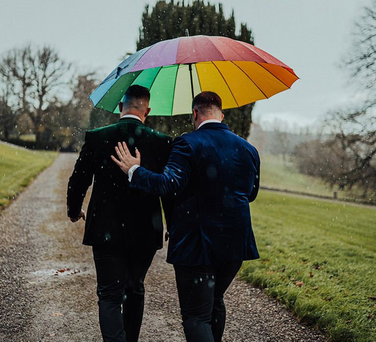 Grooms in colourful suit jackets walk under rainbow umbrella to the Castle Leslie Estate wedding venue