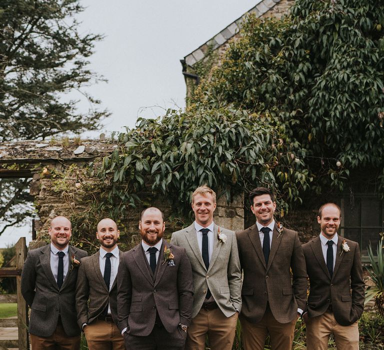 Groomsmen in different shades of brown trousers and jackets standing together with brown shoes and rose buttonholes