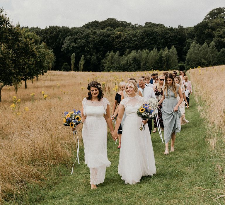 Two brides in sheer high neck wedding dresses hold hands and walk through a field as they lead their wedding guests to the barn venue