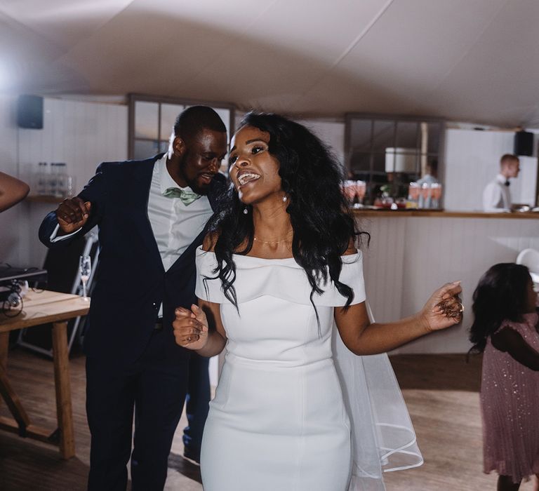 Bride in off the shoulder wedding dress with gold and pearl necklace and earrings dances with groom at reception 