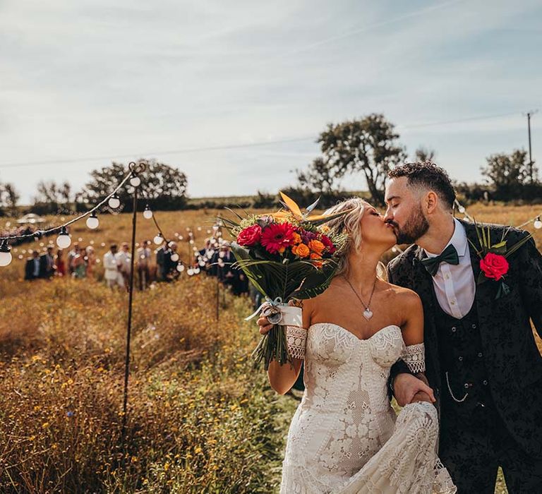 Bride in strapless lace boho wedding dress kisses groom in black patterned three piece suit and rose buttonhole as they lead guests to reception