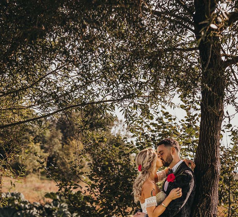 Bride and groom share a kiss against a tree with bride in strapless lace boho wedding dress and groom in black patterned suit