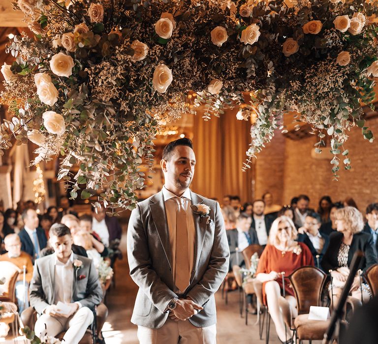 Groom in grey suit jacket with white trousers and pink tie waist patiently at the altar to see his bride