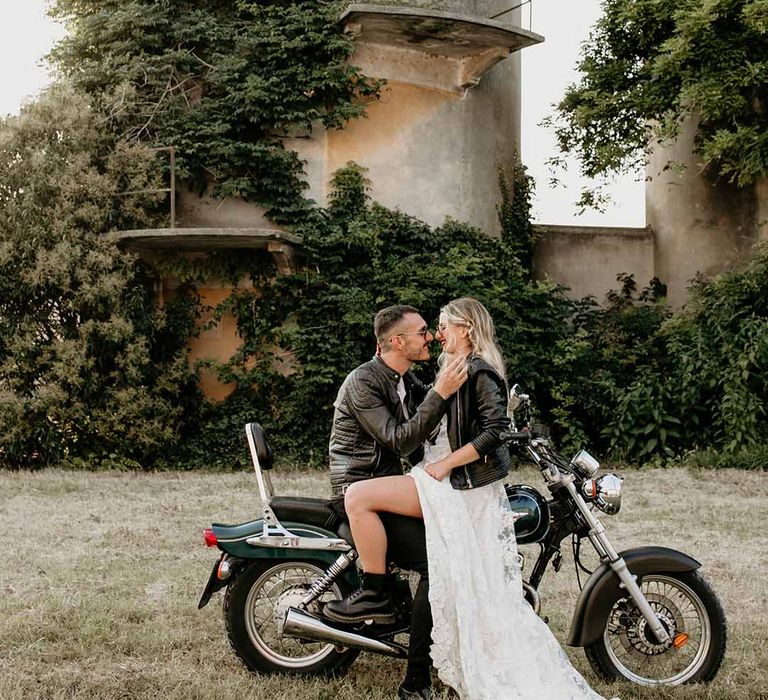 Bride in long slit wedding dress with leather jacket and boots and groom in leather jacket on dark green motorbike